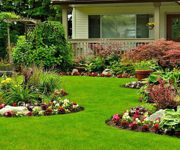 A neatly landscaped front yard featuring lush green grass, flowering garden beds with rocks, and a house with a porch.