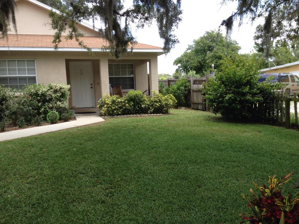A beige house with a tan roof, a green lawn, and various landscaping bushes, viewed from the front yard.