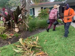 Two people in work clothes carry black pots while landscaping the front yard of a suburban house.