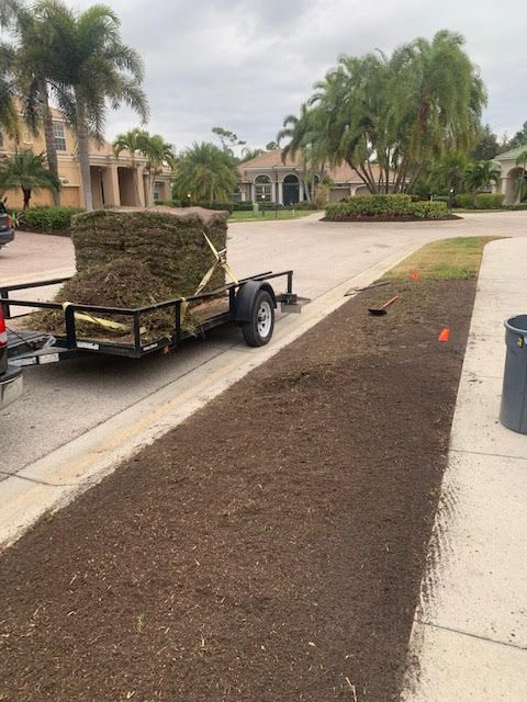 A utility trailer loaded with sod sits on a residential street next to a prepared strip of bare soil ready for landscaping.