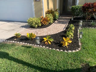 A curved brick walkway leads to a front entrance, flanked by dark mulch beds with croton plants and solar path lights.