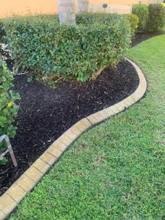 A curved border of tan stone pavers separates a dark mulch garden bed with green shrubs from a mowed green lawn.