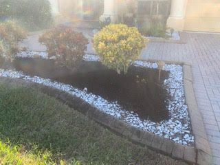 A triangular garden bed with dark mulch, two small shrubs, and a white rock border next to a paved driveway.