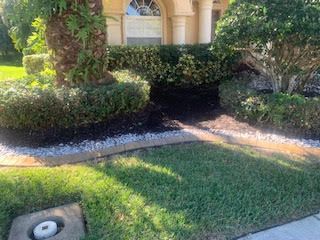 A manicured landscape features dark mulch beds, white decorative stones, a palm tree, and shrubs in front of a home.