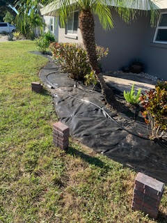 A garden bed being prepped with landscape fabric and bricks along a gray house exterior, next to a grassy lawn.
