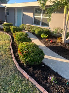 A front yard features a concrete walkway lined with dark mulch, rounded green bushes, and a red brick border.