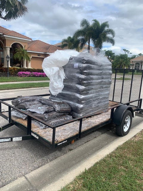 A utility trailer parked on a suburban street, loaded with a large, shrink-wrapped pallet of soil bags and loose bags.