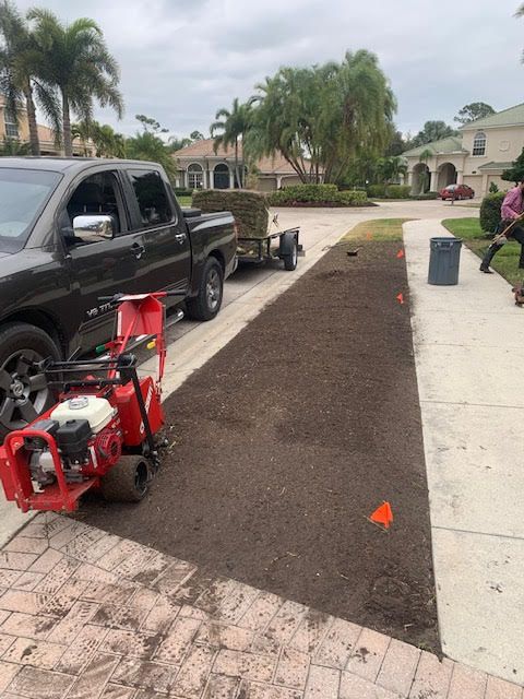 A red sod cutter sits on a strip of freshly tilled soil next to a pickup truck and trailer on a sunny residential street.