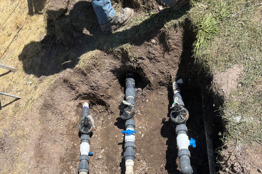 Three exposed black-and-white water pipes in a dirt trench outdoors.
