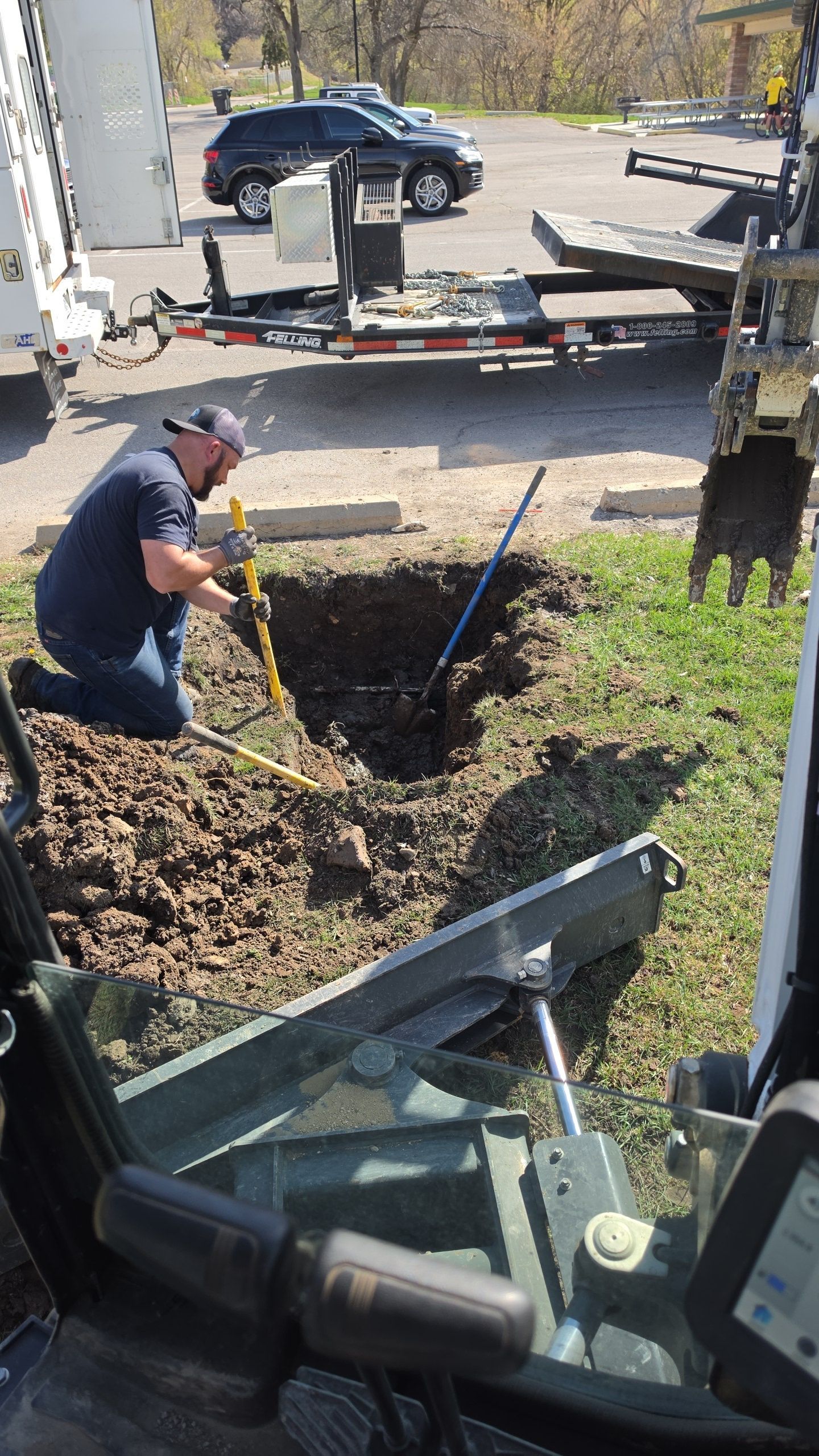 Worker digging a trench beside a trailer, seen from inside a construction vehicle.