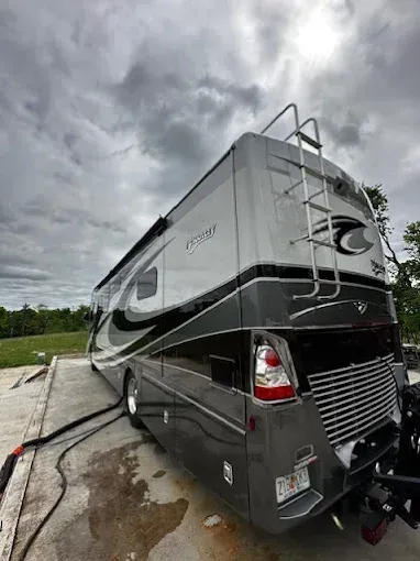 Black-and-white RV parked on wet pavement under a cloudy sky, side view with rear ladder and awning.