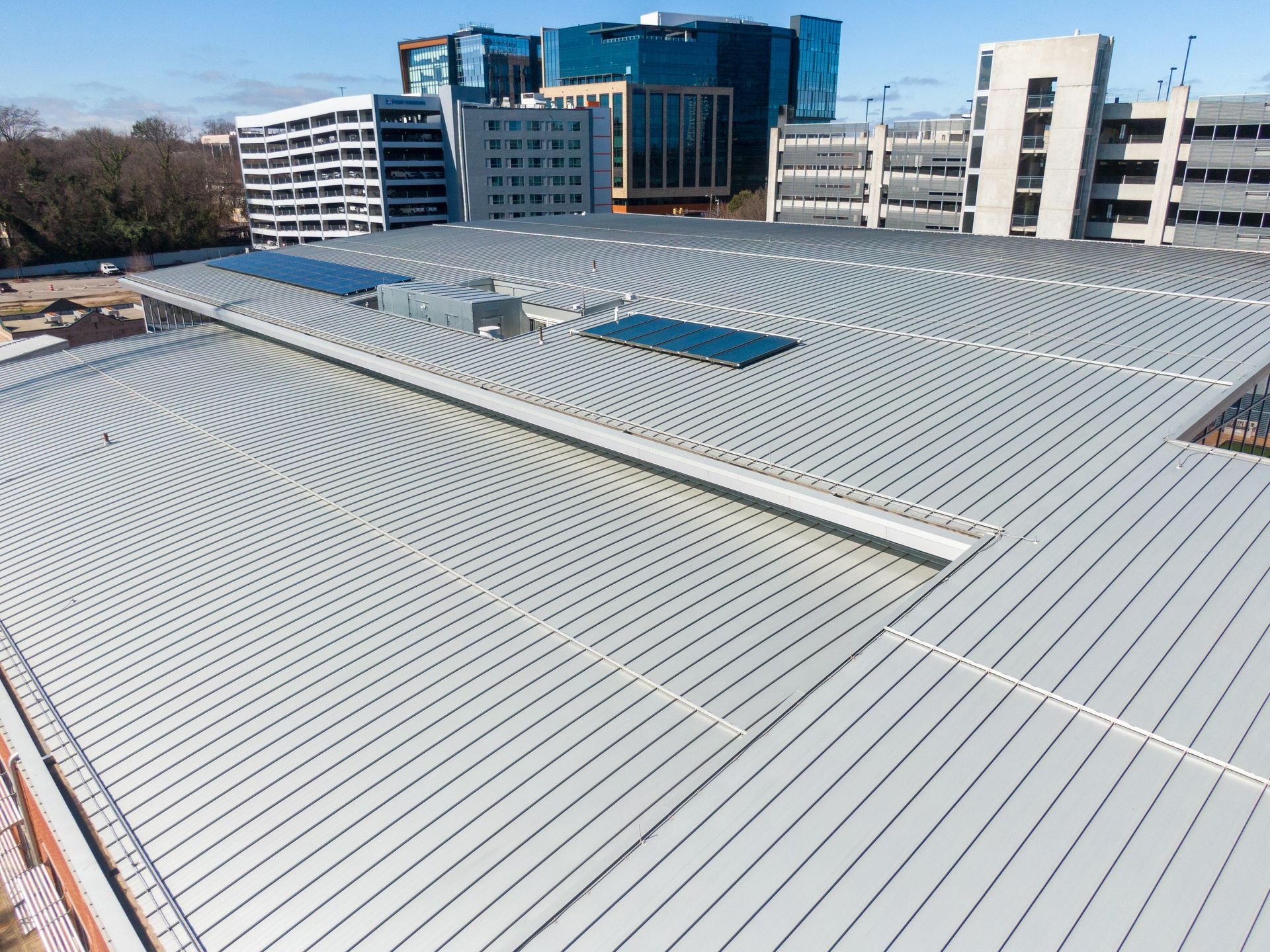 Metal roof of a modern building with several tall structures in the background on a sunny day.