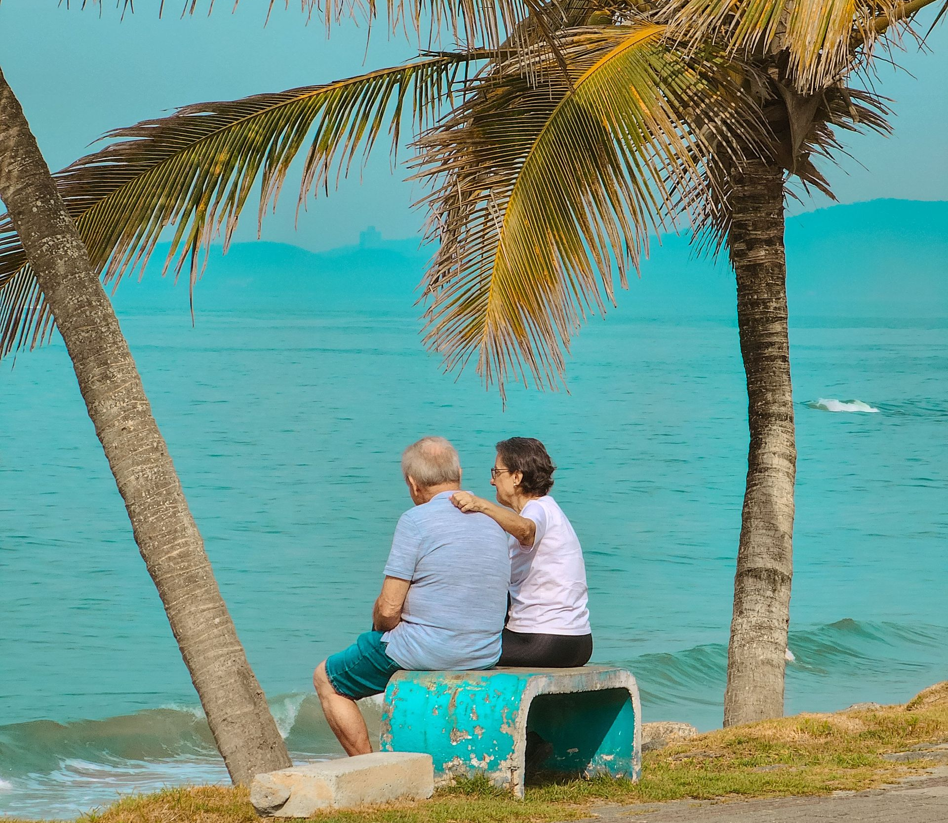 Two people sit on a bench by turquoise water under palm trees, facing the sea.