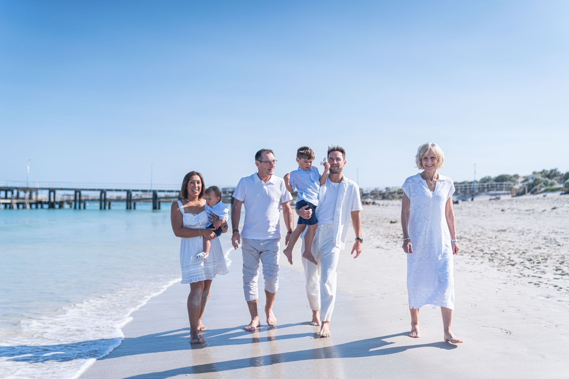 Five people walking barefoot on a bright beach near the water with a pier in the background