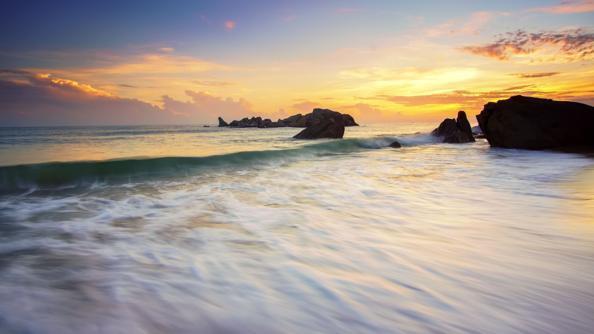 Sunset over a calm beach with waves, rocky outcrops, and orange clouds in the sky
