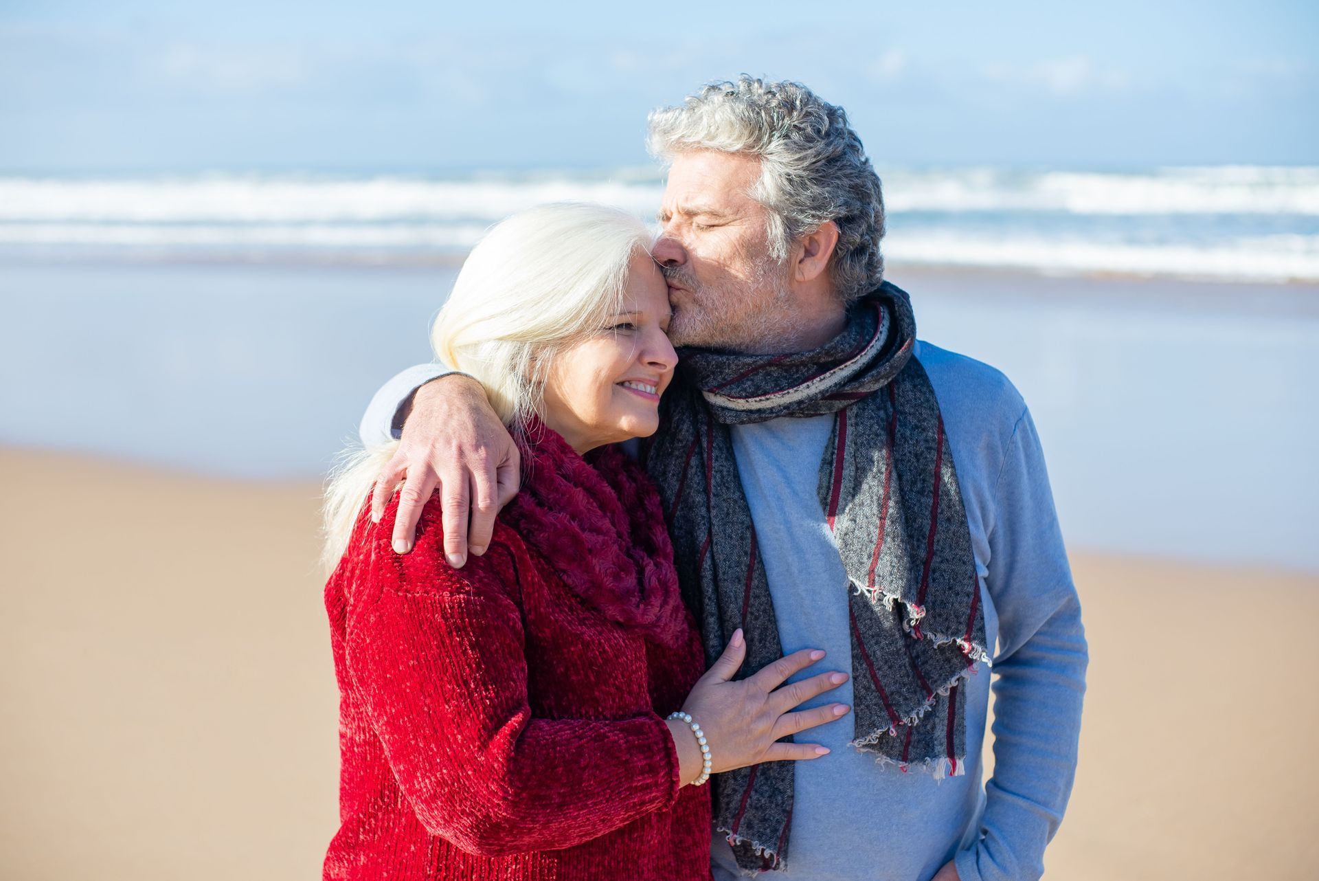 Couple embracing on a beach, wrapped in winter coats and scarves, with waves in the background