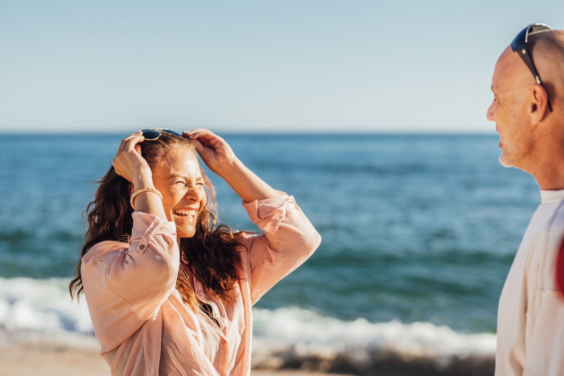 Two people smiling and talking on a sunny beach with the ocean behind them