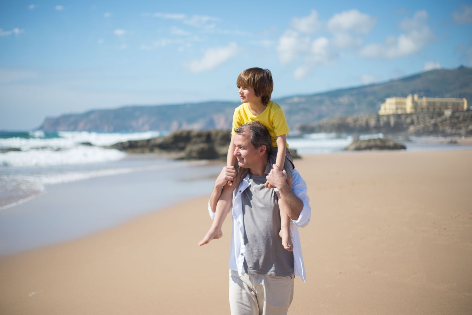 Adult carrying child on shoulders on a sunny beach with waves and cliffs in the background
