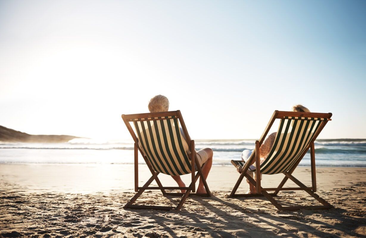 Two people in wooden chairs relax on a sunny beach at sunrise over calm water