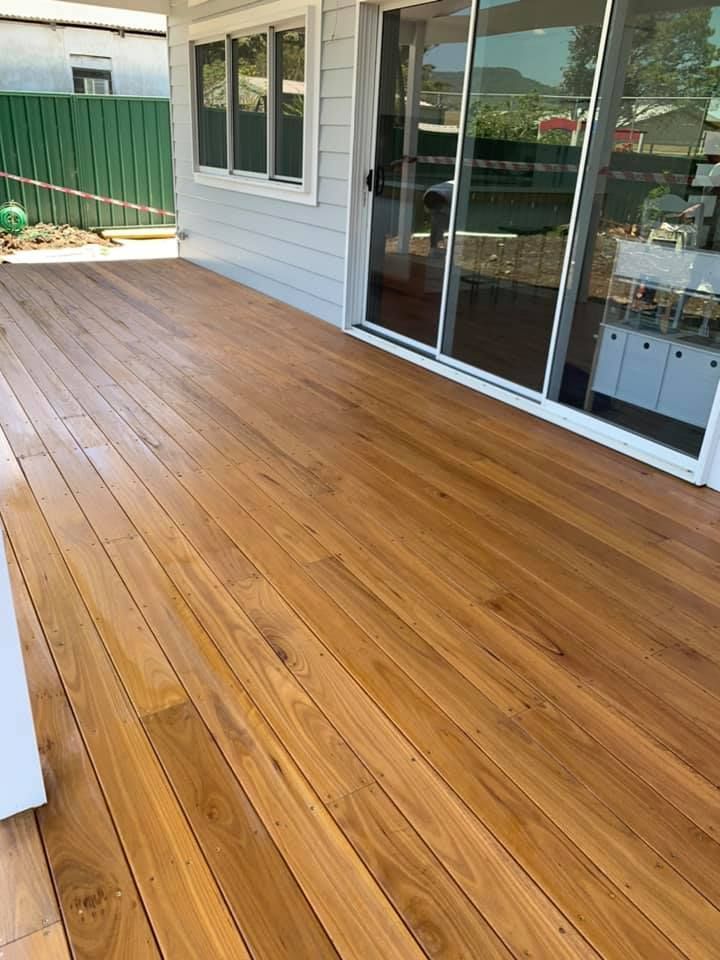Wooden deck extending from a light gray house with large glass sliding doors; green fence in background.