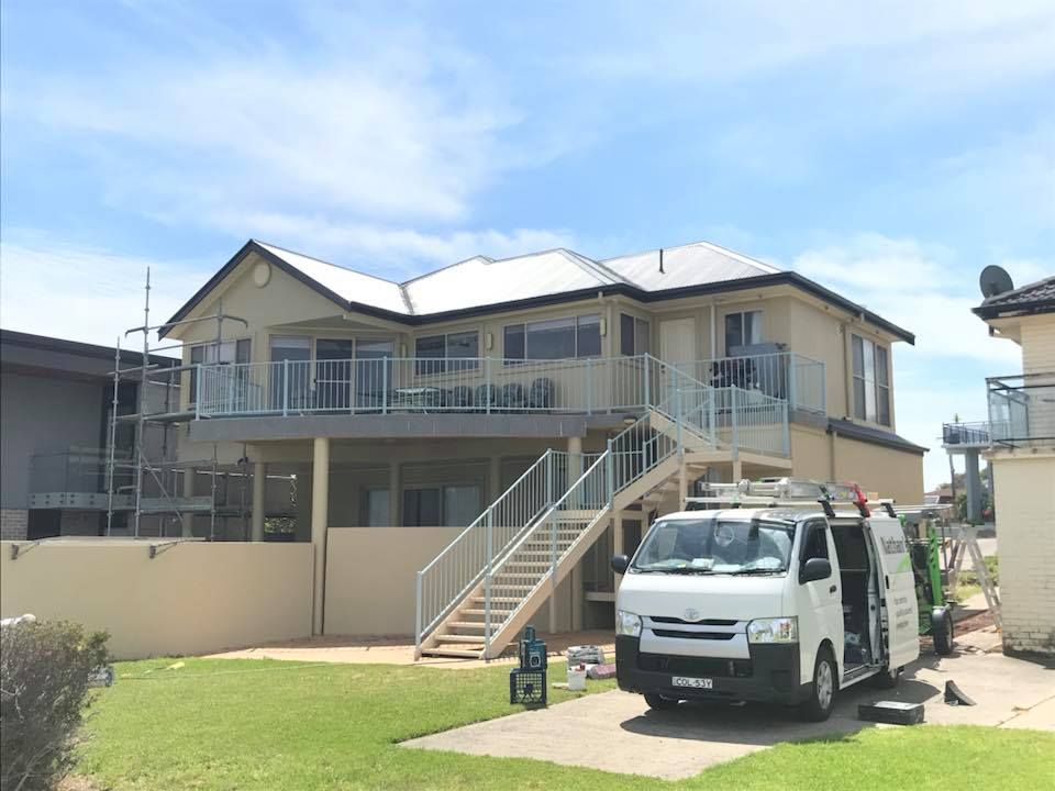 Two-story house with a white van in front. White railings, beige siding, blue sky.