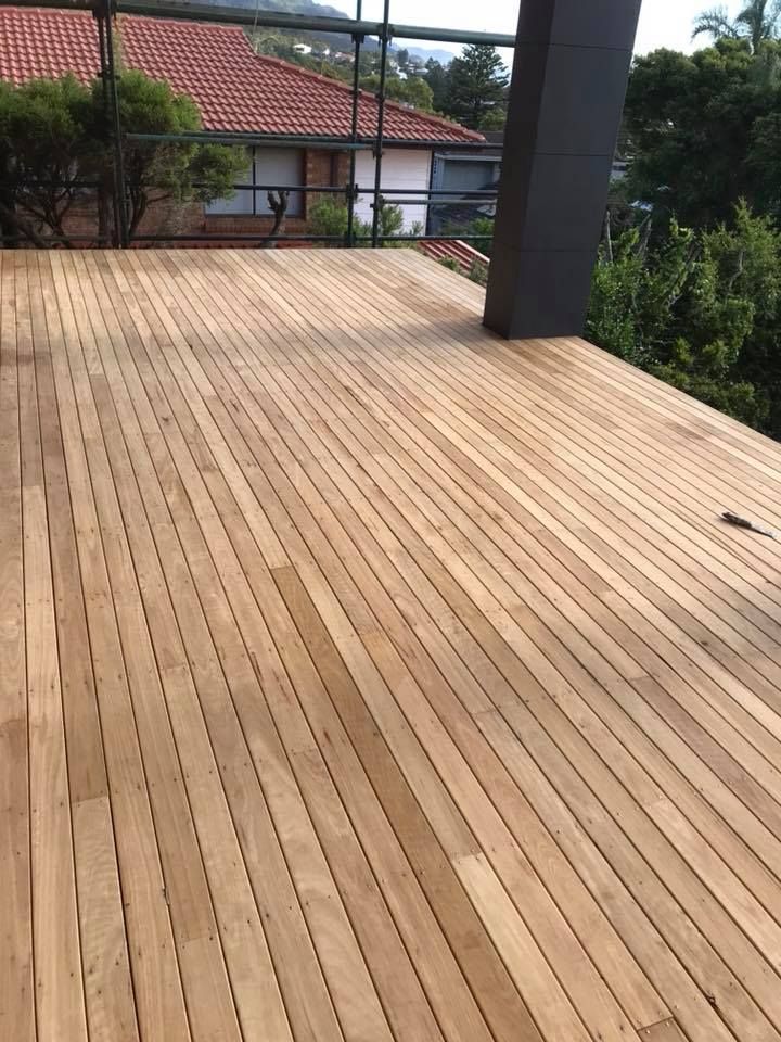 Wooden deck with light-colored, horizontal planks. In the background, a house with a red tiled roof is visible.