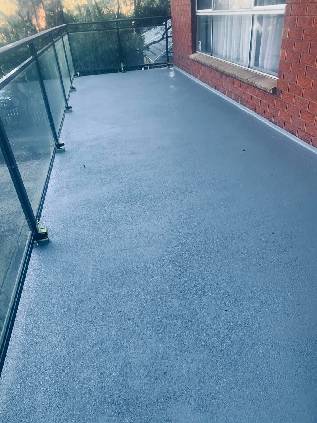 A balcony with a gray, textured surface, glass railing, and brick wall.