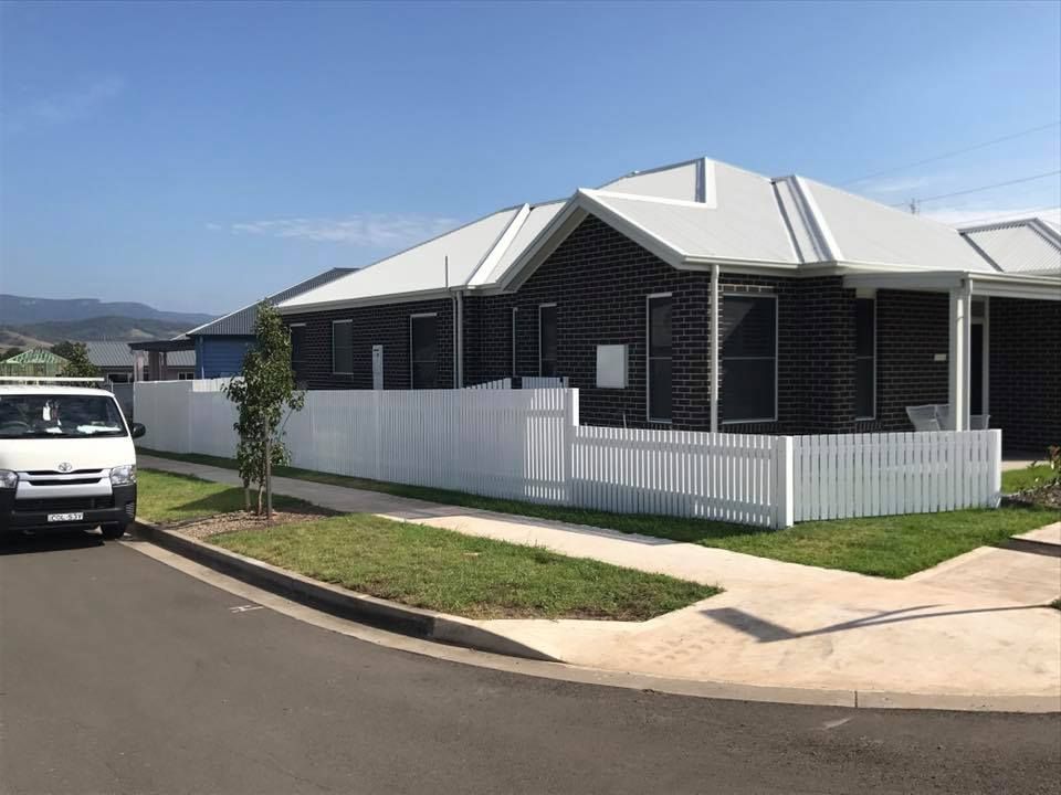 White picket fence surrounds a modern brick house with a white roof on a sunny day. A van is parked on the street.