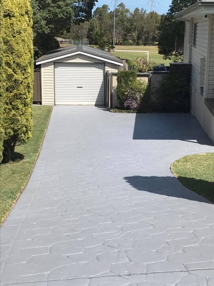Gray patterned driveway leading to a garage and house with surrounding grass and trees under a sunny sky.