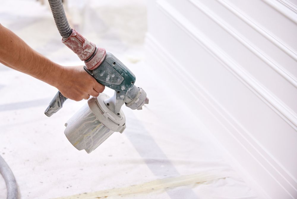 Person spray-painting white trim with a paint sprayer, on a covered floor.