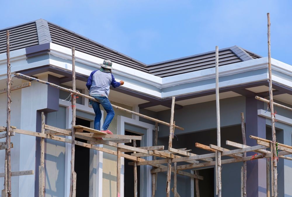 Construction worker on scaffolding, painting a house exterior, blue sky background.