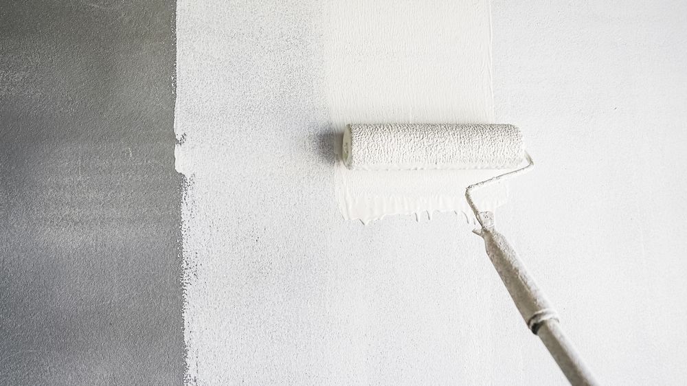 Paint roller applying white paint to a wall, covering a gray section.