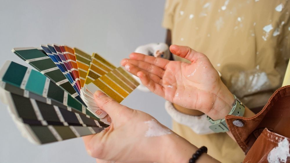 Person holding a paint swatch fan, comparing colors with another person wearing paint-splattered clothing.