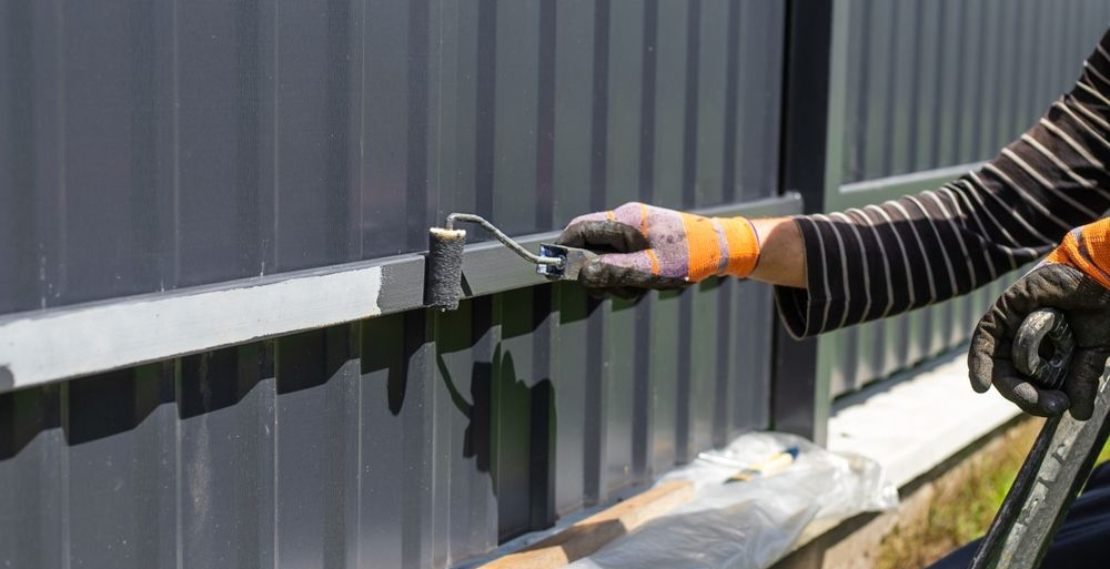 Man in safety glasses spray paints a brown metal fence outdoors.