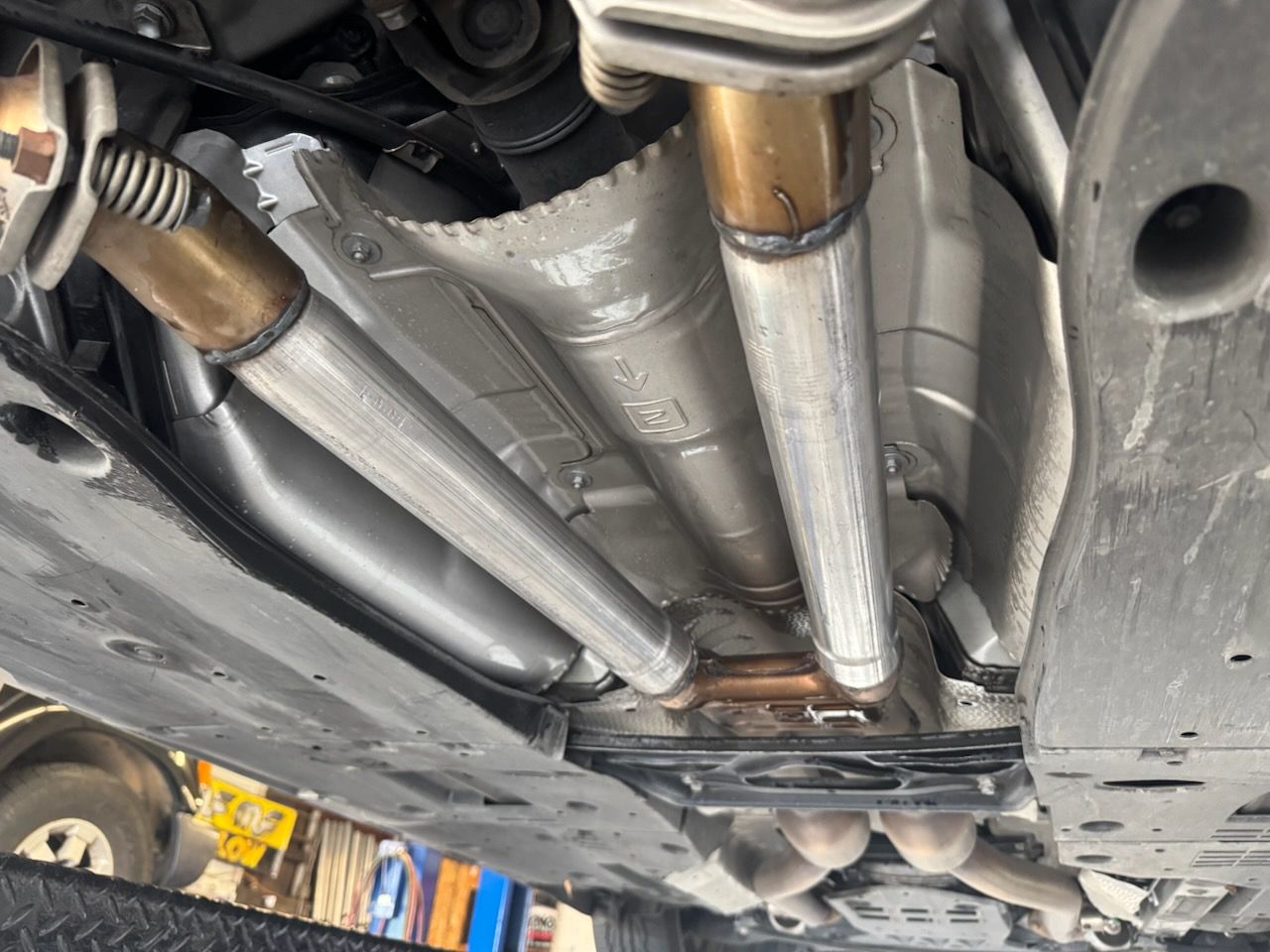 An underside view of a vehicle showing a silver dual exhaust system, metallic heat shields, and a dark chassis.