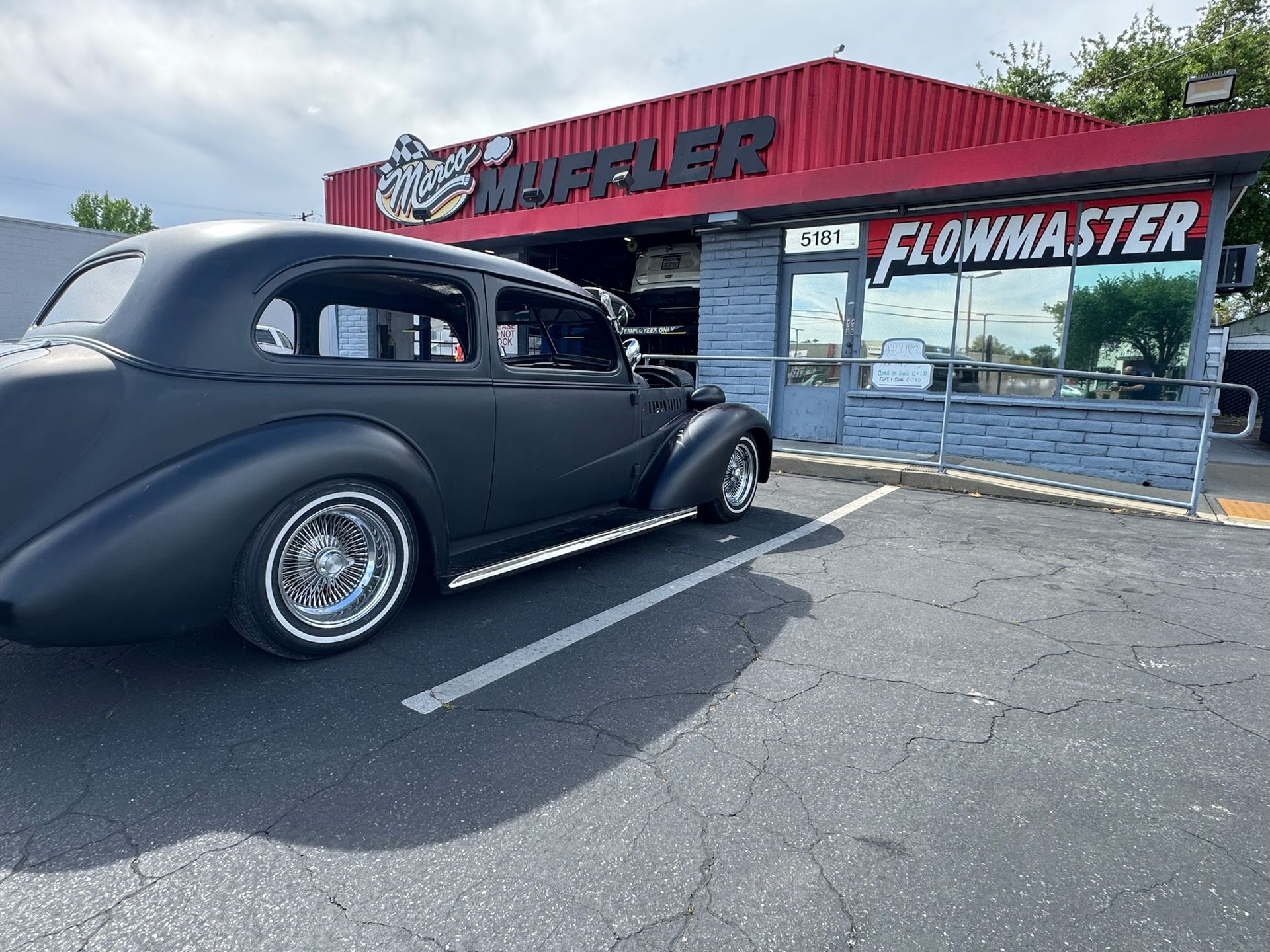 A matte black vintage car parked in front of a red-roofed muffler shop with a 