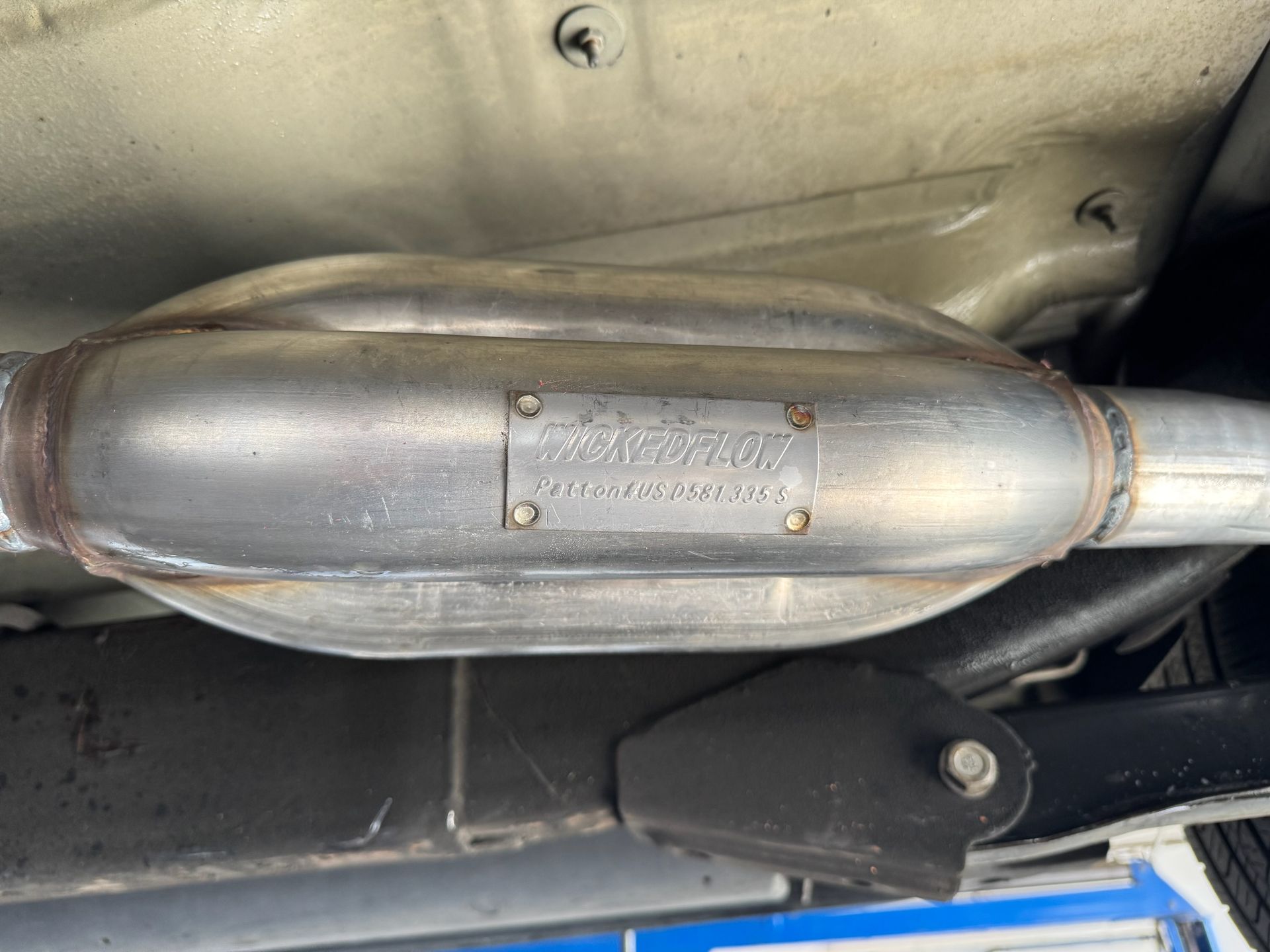 A metal vehicle muffler with a riveted Flowmaster brand plaque visible under a car chassis.