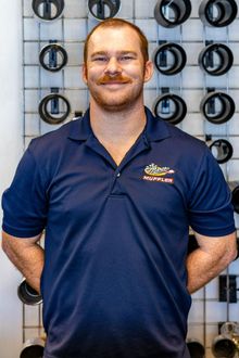 A smiling person wearing a blue polo shirt with a logo, standing in front of a grid wall displaying hardware supplies.