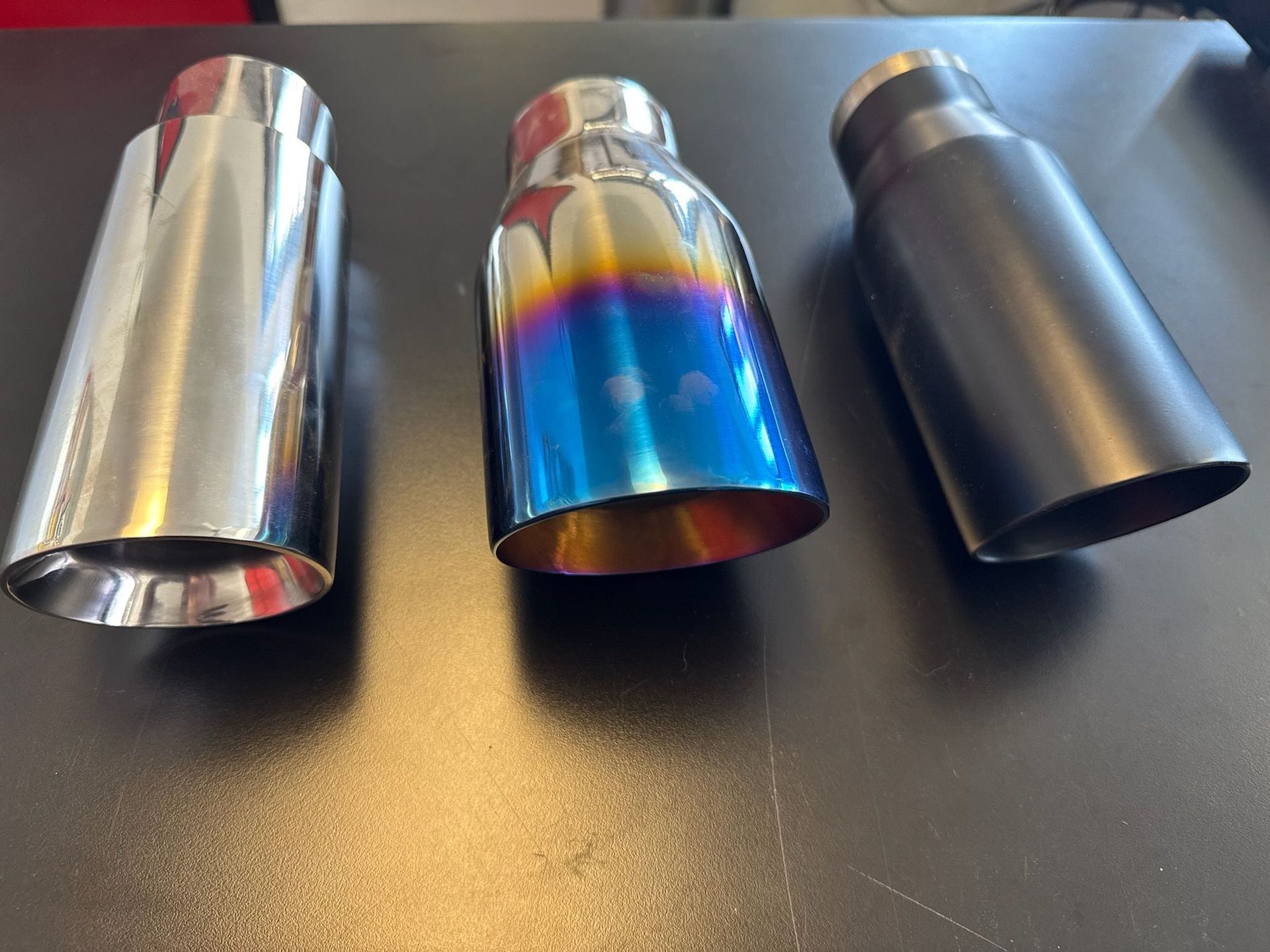 Three car exhaust tips in chrome, rainbow-colored titanium, and matte black finishes sitting on a dark surface.