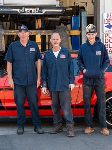 Three people in work uniforms stand in front of a red car in a garage with a vehicle on a lift in the background.
