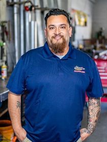 A person with a beard and tattoos on their arms wearing a navy blue polo shirt, standing in a workshop setting.
