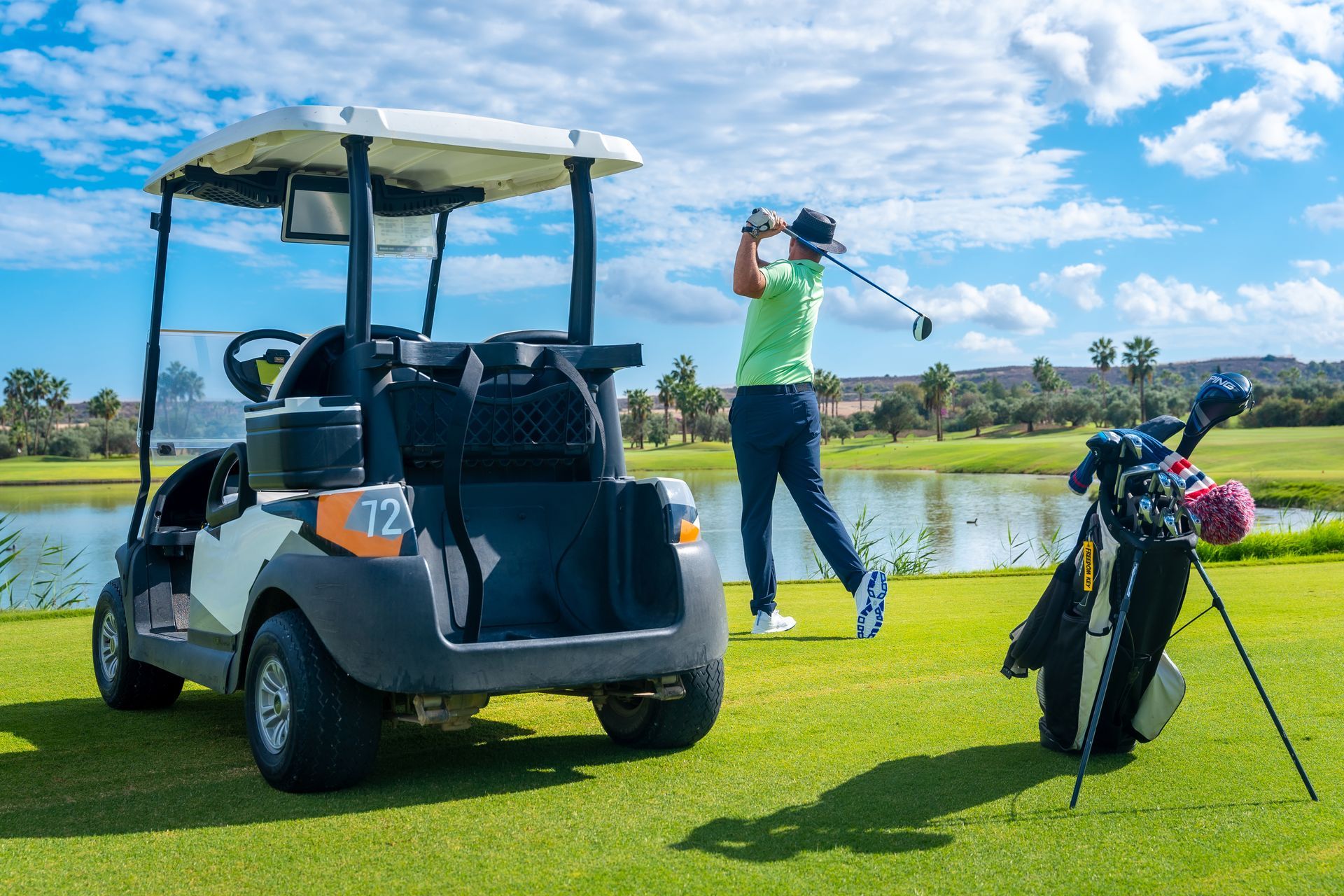 Golfer in green shirt swinging on a golf course beside a golf cart and clubs by a pond