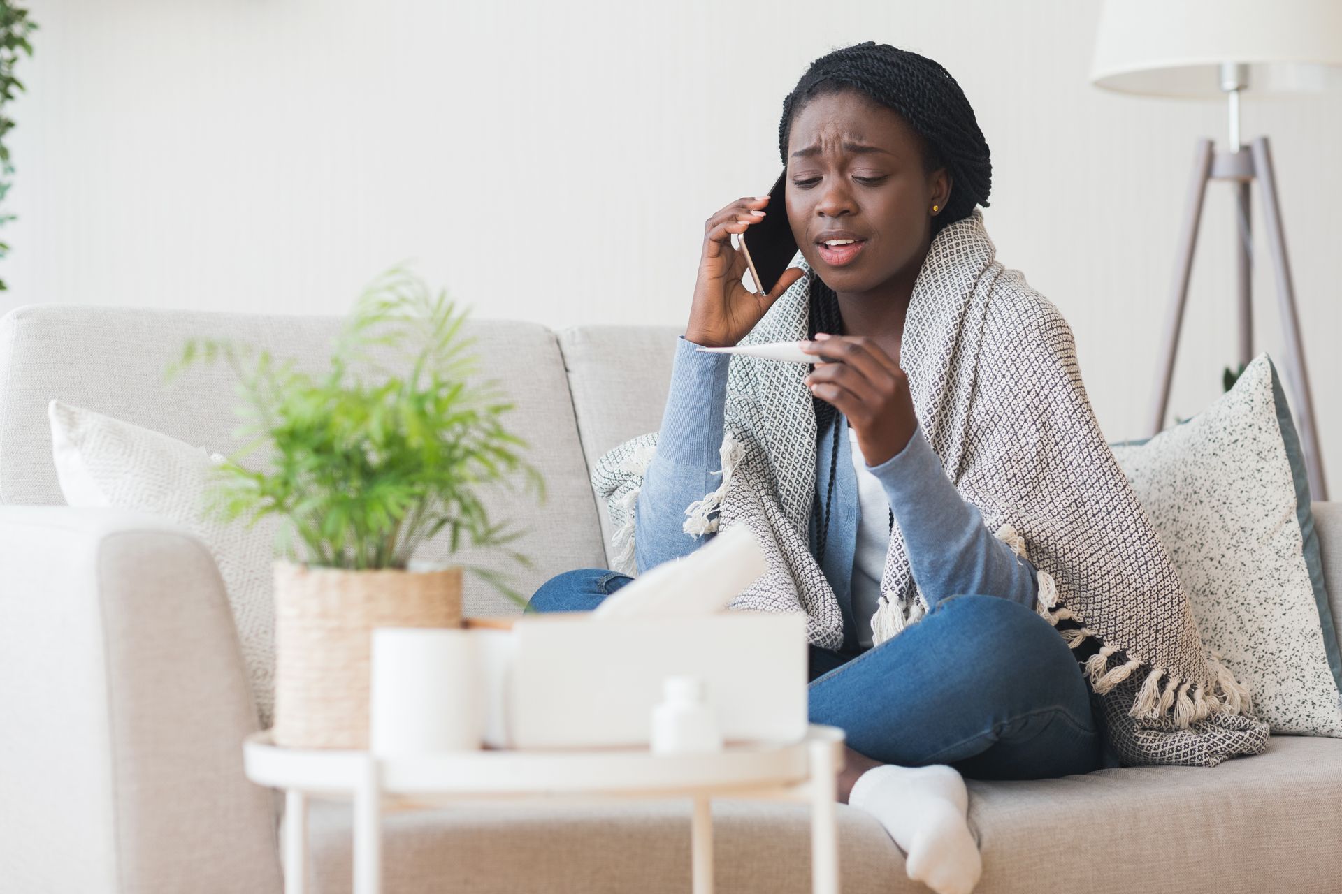 Woman sitting on a couch, talking on a phone and holding a tissue, looking distressed in a bright living room