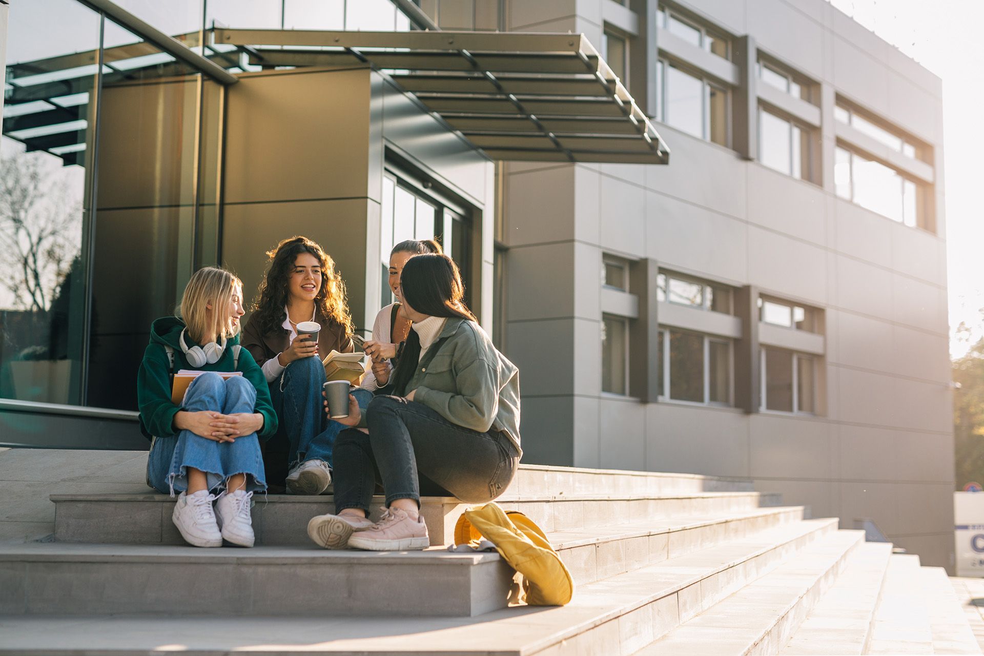Three students sit chatting on steps outside a modern school building.