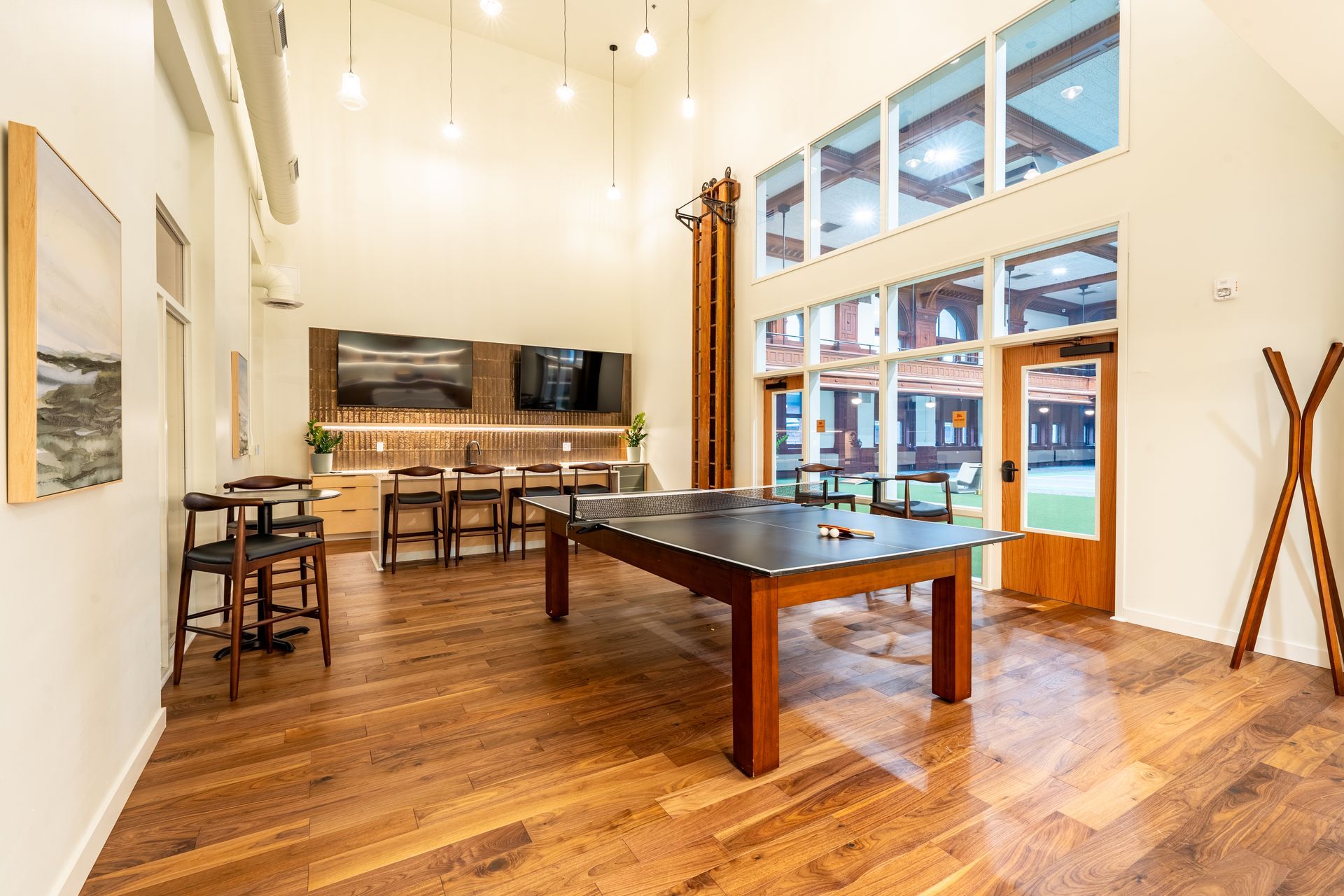 Interior of a lounge with a pool table, bar seating, TVs, and large windows. Wooden floors and white walls.