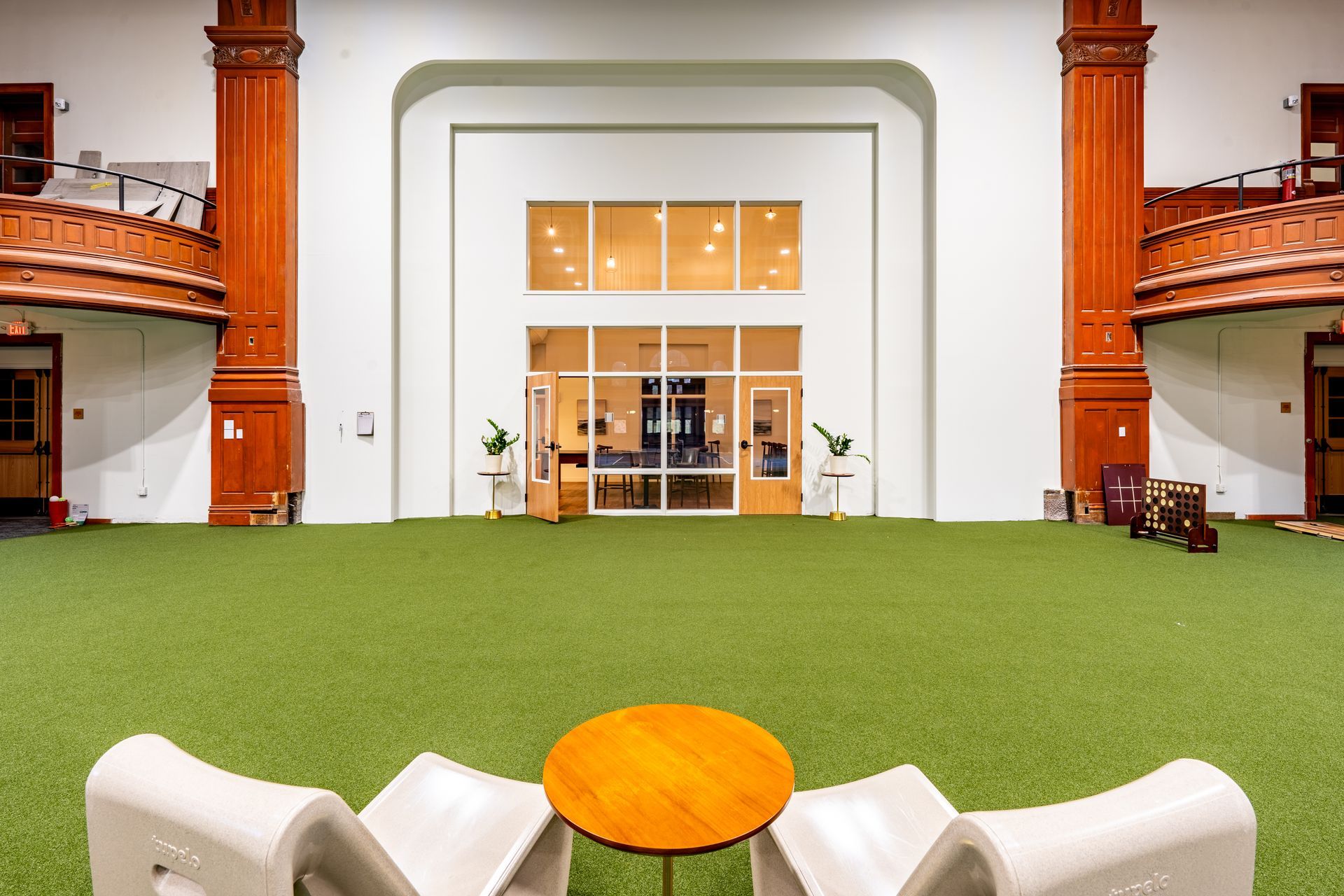 Open space with green flooring, white chairs, a round table, and a glass-paneled doorway. Red columns flank the sides.