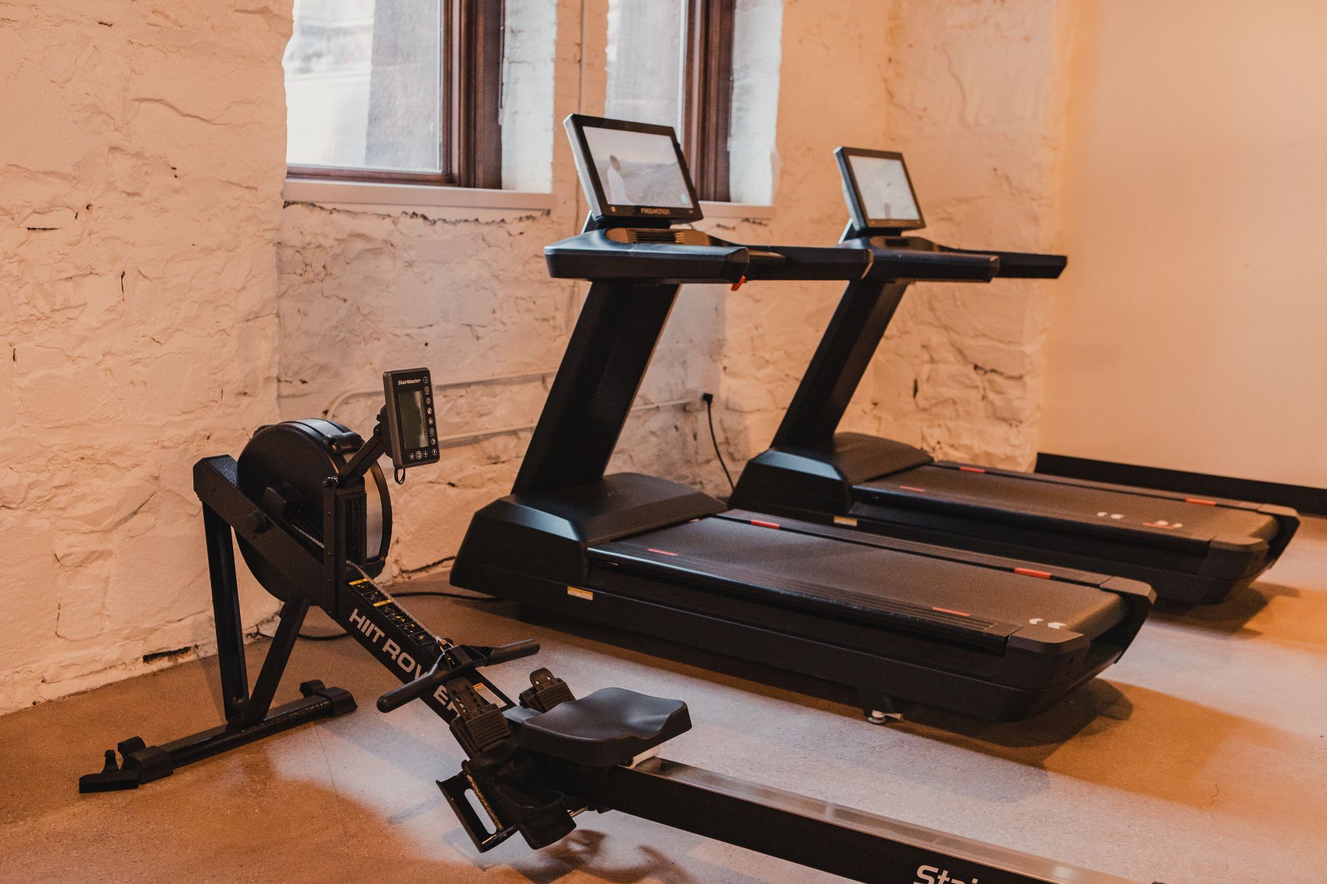 Rowing machine and two treadmills in a room with light-colored stone walls.
