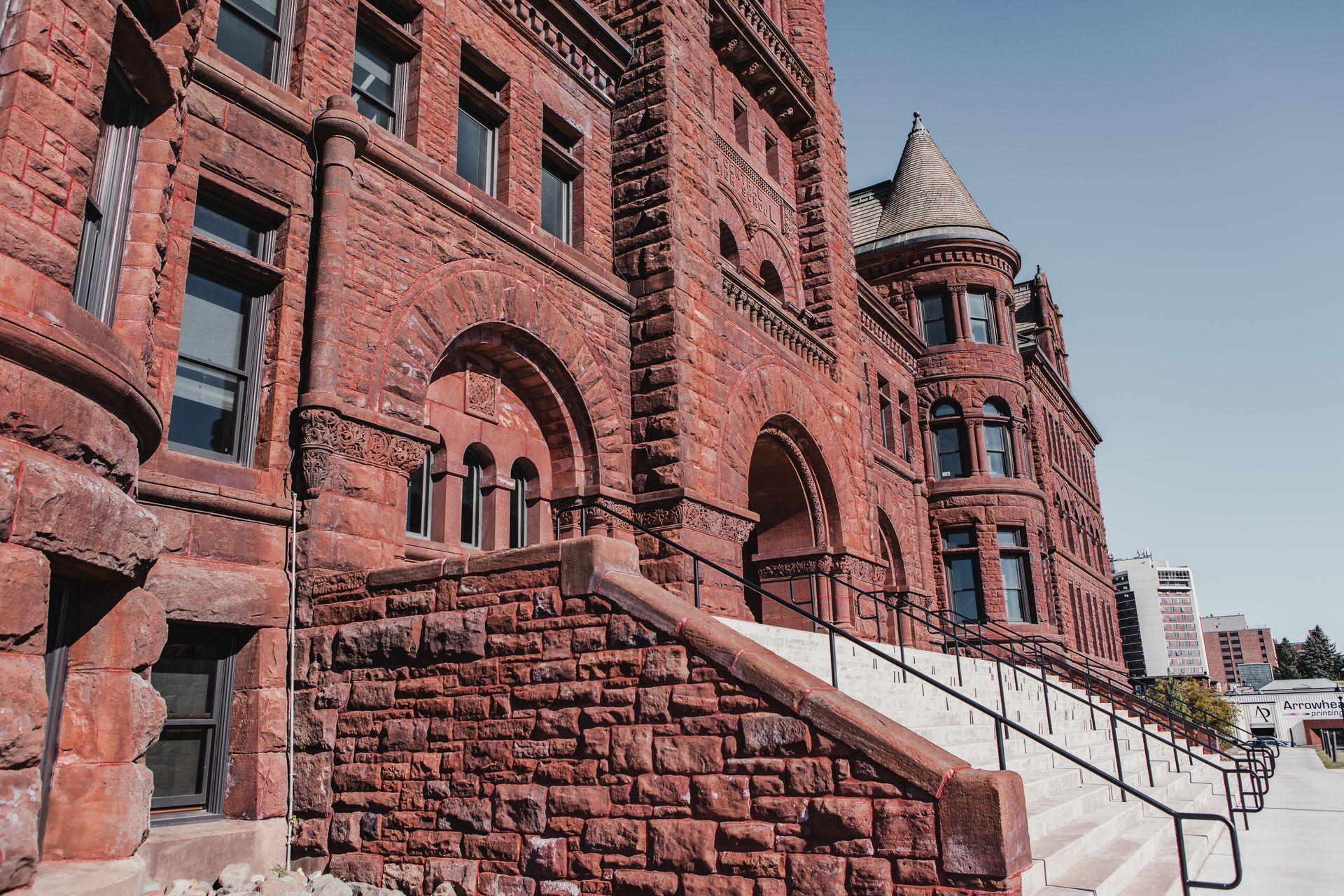 Red brick building with arched windows, steps, and a turret on a sunny day.