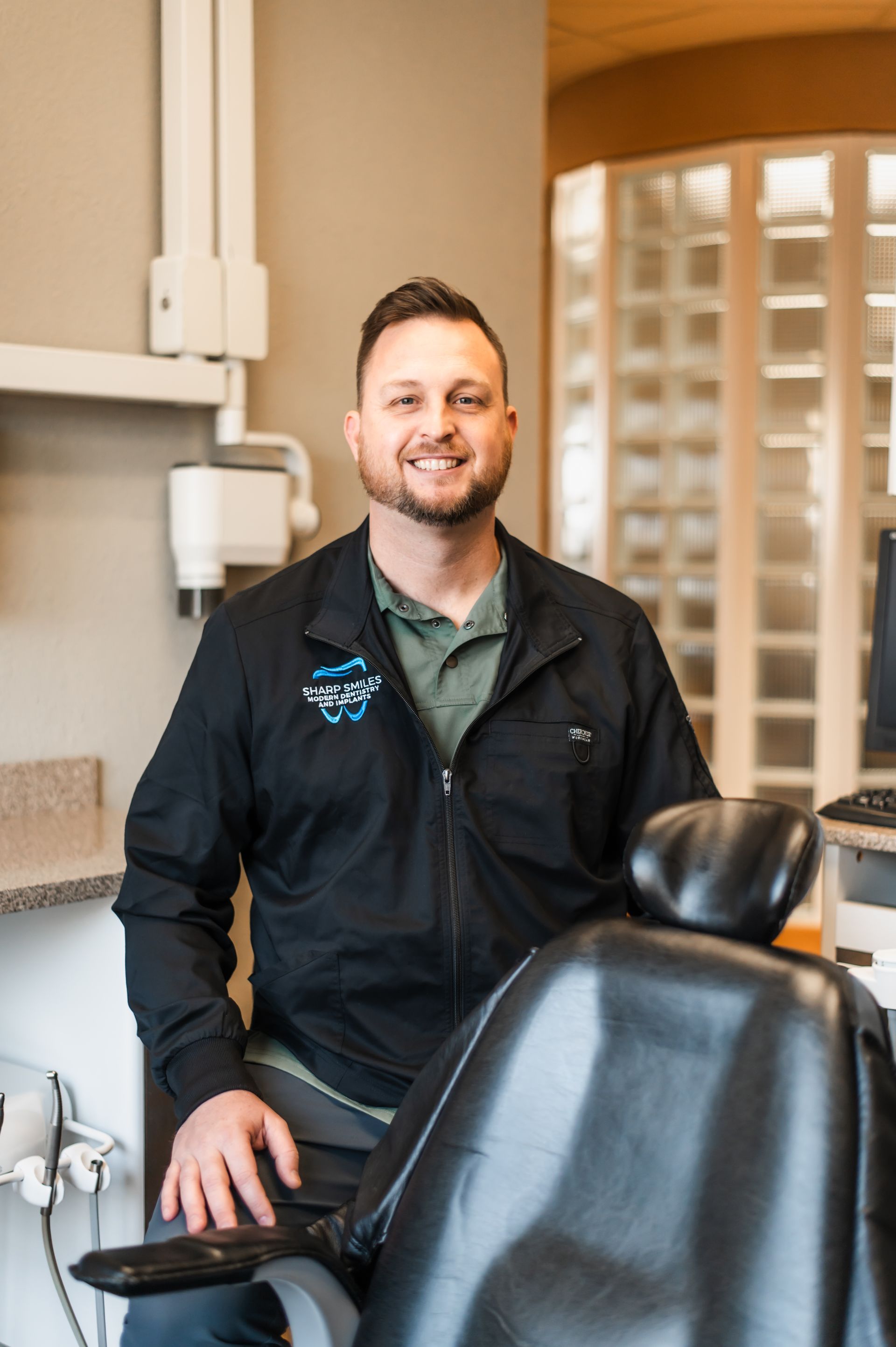 Dentist standing beside a dental chair in a bright clinic, smiling at the camera.