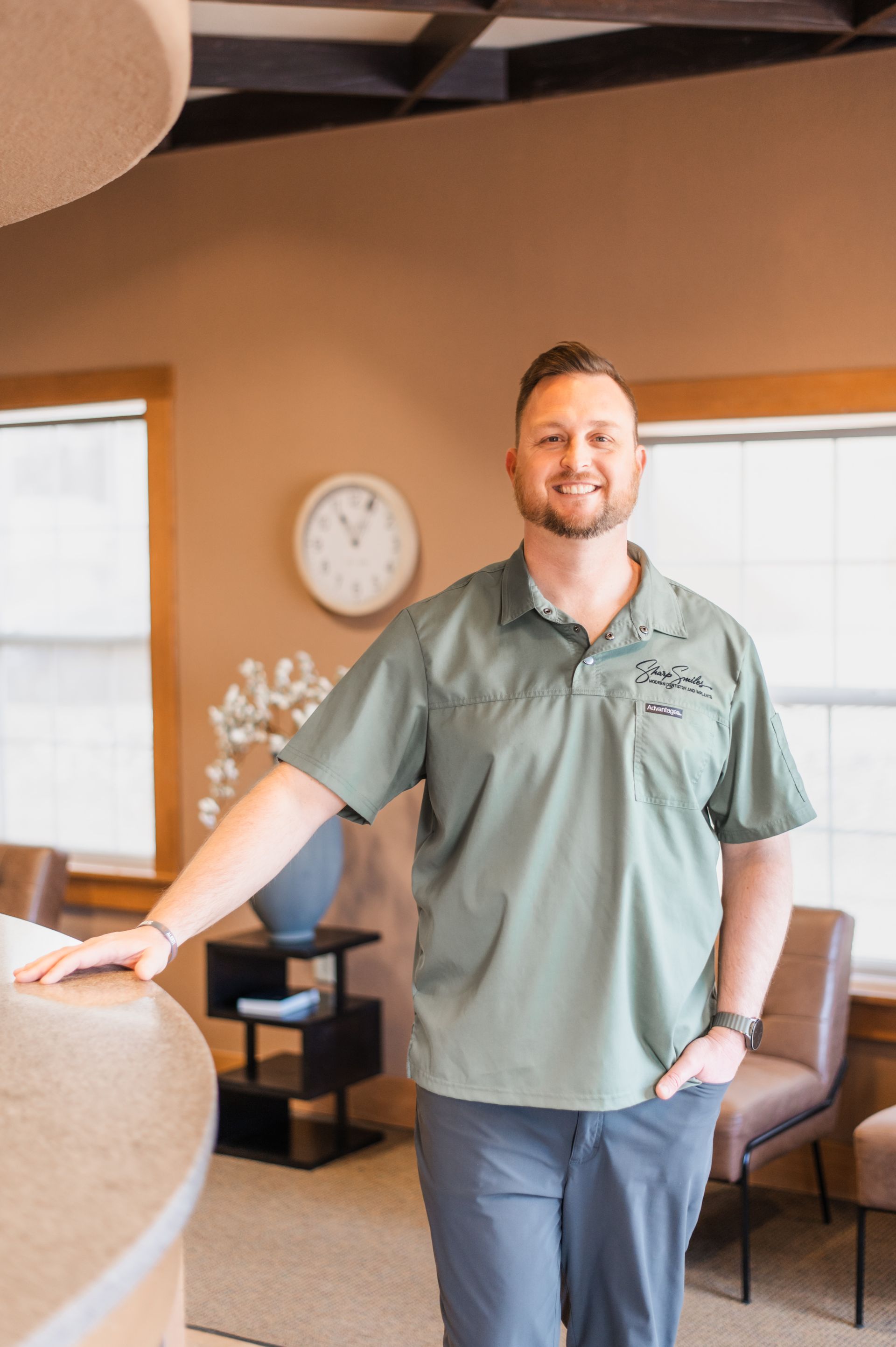 Smiling man in a green polo shirt standing in a warmly lit office lobby with chairs and a wall clock